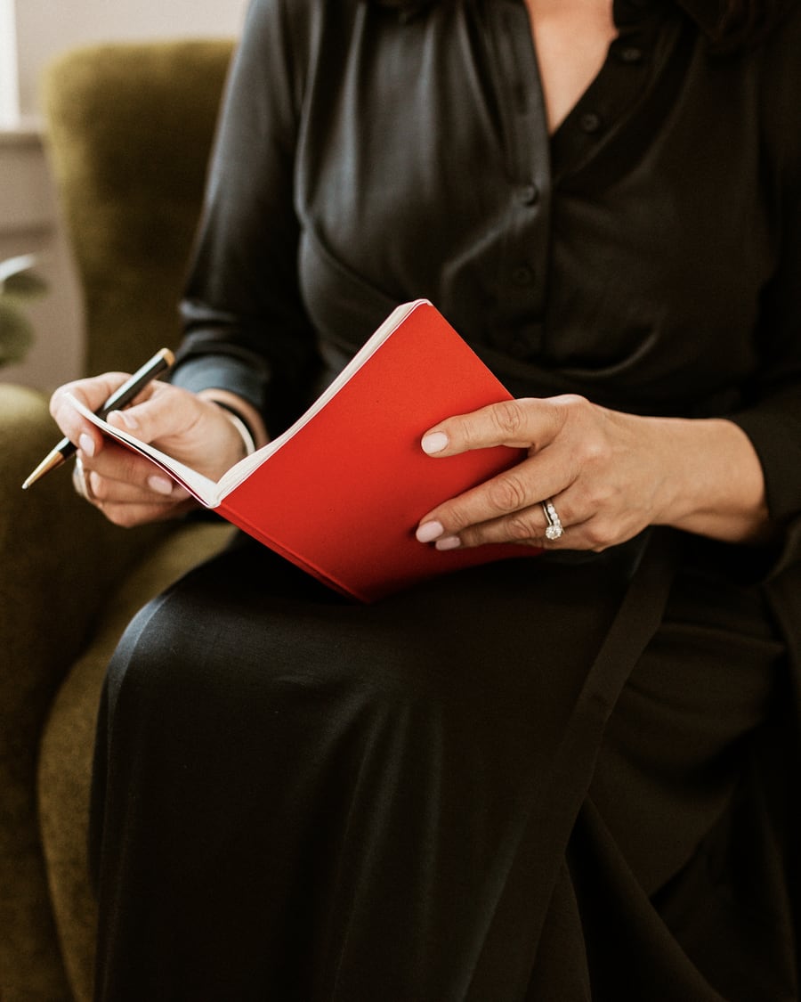 Ellen seated in an armchair, pen in hand, with a red notebook open on her lap